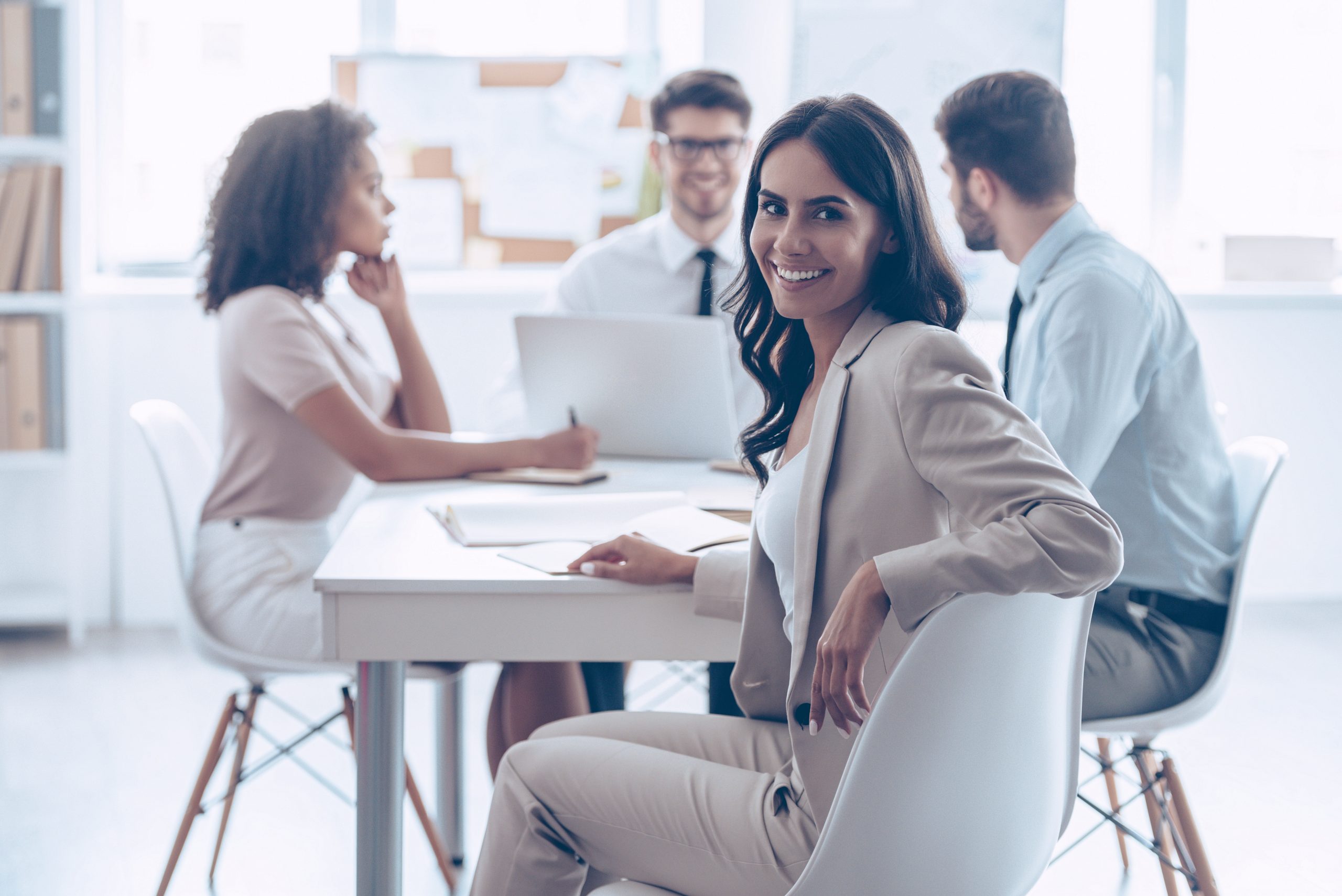 Feliz por estar na equipe. Mulher bonita e alegre olhando para a câmera com um sorriso enquanto está sentada à mesa do escritório com seus colegas de trabalho