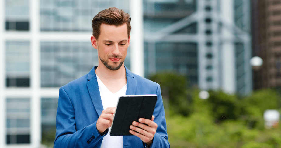 Homem em primeiro plano segura um tablet. Ao fundo prédios e árvores.