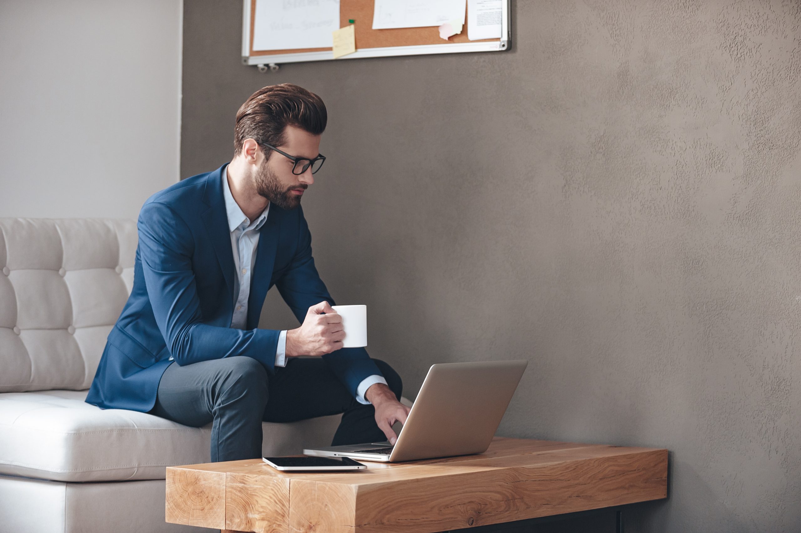 Começando um novo dia de trabalho. Jovem bonito usando óculos, segurando uma xícara de café e trabalhando com um laptop enquanto está sentado no sofá no escritório